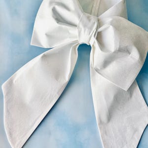 May include: A large, white bow made of fabric, with long, flowing tails. The bow is centered and appears to be handmade, with a textured surface. The background is a light blue, creating a contrast with the bright white bow.