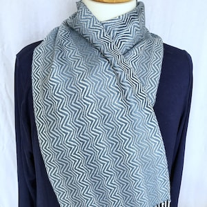 Puede incluir: Una bufanda de patrón de chevron azul y blanco con flecos. La bufanda se muestra en un maniquí con una camisa azul marino.