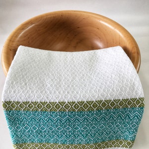 May include: A white kitchen towel with a teal and green geometric pattern border. The towel is folded and placed inside a wooden bowl.