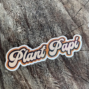 May include: A sticker with the text "Plant Papi" in a retro font style. The text is in a light brown color with a dark brown outline. The sticker is on a brown wooden surface.