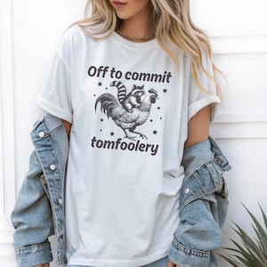 May include: White t-shirt with a graphic of a raccoon riding a rooster, surrounded by stars, and the text "Off to commit tomfoolery". The shirt has short sleeves and a relaxed fit.
