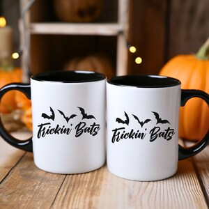 K&ouml;nnte beinhalten: Zwei wei&szlig;e Kaffeetassen mit schwarzen R&auml;ndern und Griffen. Die Tassen haben ein schwarzes Motiv mit Flederm&auml;usen und dem Text "Frickin' Bats" darauf.