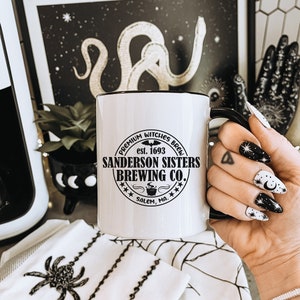 May include: White ceramic mug with a black handle and a black graphic that reads "PREMIUM WITCHES BREW est. 1693 SANDERSON SISTERS BREWING CO. *** SALEM, MA. ✱✱"