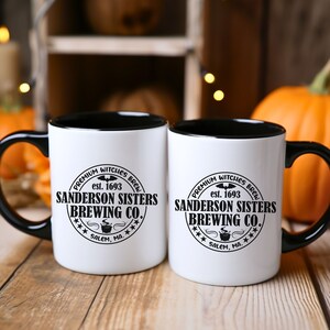 May include: Two white ceramic mugs with black handles and black rims. Both mugs feature a black and white logo that reads "Premium Witches Brew est. 1693 Sanderson Sisters Brewing Co. *** Salem, MA. ***"