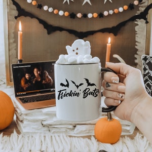 K&ouml;nnte beinhalten: Eine wei&szlig;e Keramiktasse mit schwarzem Text, der "Frickin Bats" lautet, und einem schwarzen Fledermaus-Silhouetten-Design. Die Tasse ist gef&uuml;llt mit hei&szlig;er Schokolade, die mit Marshmallows in Form eines Geistes gekr&ouml;nt ist.