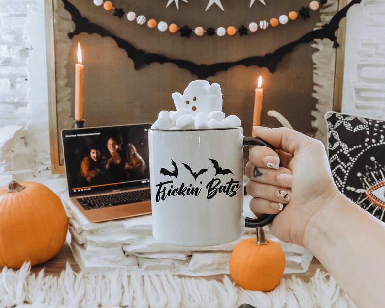 K&ouml;nnte beinhalten: Eine wei&szlig;e Keramiktasse mit schwarzem Text, der "Frickin Bats" lautet, und einem schwarzen Fledermaus-Silhouetten-Design. Die Tasse ist gef&uuml;llt mit hei&szlig;er Schokolade, die mit Marshmallows in Form eines Geistes gekr&ouml;nt ist.