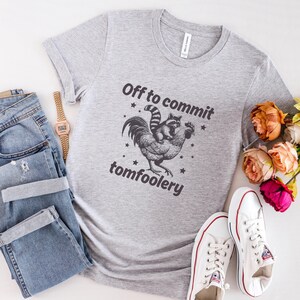 May include: A light gray t-shirt with the text "Off to commit tomfoolery" above and below a graphic of a raccoon riding a rooster. The shirt is paired with blue jeans, a watch, and white sneakers.