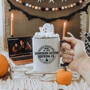 May include: White ceramic mug with a black and white graphic of a witch's cauldron and the text "PREMIUM WITCHES BREW est. 1693 SANDERSON SISTERS BREWING CO. **+ SALEM, MA. ***". The mug is filled with marshmallows and a ghost-shaped marshmallow on top.