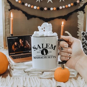 May include: White ceramic mug with black text that reads "Salem Est. 1626 Massachusetts" and a black silhouette of a witch. The mug is filled with hot chocolate topped with marshmallows and a ghost-shaped marshmallow.