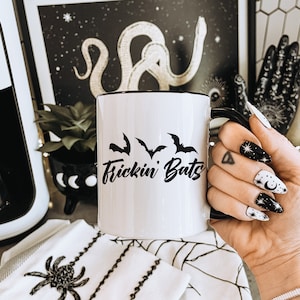 K&ouml;nnte beinhalten: Eine wei&szlig;e Keramiktasse mit schwarzem Rand und Henkel. Die Tasse hat ein schwarzes Motiv mit Flederm&auml;usen und dem Text "Frickin Bats".