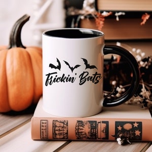 K&ouml;nnte beinhalten: Wei&szlig;e Keramiktasse mit schwarzem Rand und Henkel. Die Tasse hat ein schwarzes Motiv mit Flederm&auml;usen und dem Text "Frickin' Bats".