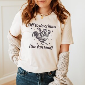May include: Cream-colored t-shirt featuring a graphic of a raccoon riding a rooster, with the text "Off to do crimes (the fun kind)". The design is in black and white, with small star accents. The shirt is made of a soft material.
