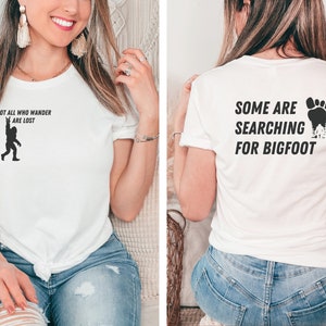 May include: White t-shirt with a black silhouette of Bigfoot on the front and the text "Not all who wander are lost". The back of the shirt has the text "Some are searching for Bigfoot" with a black footprint and trees.