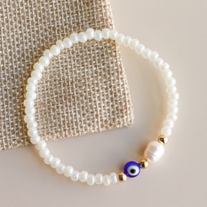 Peut inclure: Un bracelet de perles compos&eacute; de petites perles en verre blanc, d'une perle d'eau douce blanc cass&eacute;, d'une perle en verre bleu et blanc en forme d'&oelig;il protecteur et de deux minuscules perles intercalaires m&eacute;talliques dor&eacute;es.