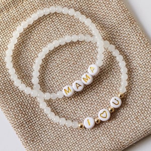 May include: Two white beaded bracelets with gold lettering. One bracelet spells "MAMA" and the other spells "I LOVE U" with a heart in place of the "O".