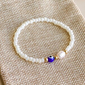 Peut inclure: Un bracelet de perles compos&eacute; de petites perles blanches, d'une perle en verre bleu et blanc en forme d'&oelig;il protecteur, d'une perle d'eau douce irr&eacute;guli&egrave;re et de petites perles intercalaires m&eacute;talliques dor&eacute;es dispos&eacute;es en cercle.
