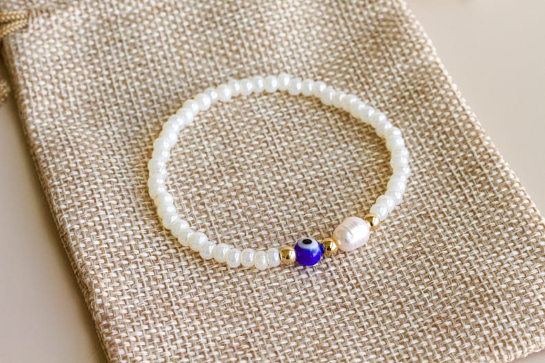 Peut inclure: Un bracelet de perles compos&eacute; de petites perles blanches, d'une perle en verre bleu et blanc en forme d'&oelig;il protecteur, d'une perle d'eau douce irr&eacute;guli&egrave;re et de petites perles intercalaires m&eacute;talliques dor&eacute;es dispos&eacute;es en cercle.