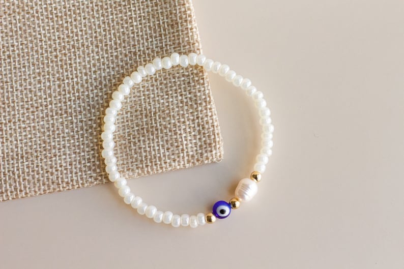 Peut inclure: Un bracelet de perles compos&eacute; de petites perles en verre blanc, d'une perle d'eau douce blanc cass&eacute;, d'une perle en verre bleu et blanc en forme d'&oelig;il protecteur et de deux minuscules perles intercalaires m&eacute;talliques dor&eacute;es.