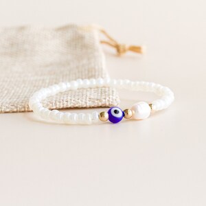 Peut inclure: Un bracelet compos&eacute; de petites perles blanches opaques, d'une perle unique en forme d'&oelig;il bleu et blanc, d'une petite perle d'eau douce blanche et de deux minuscules perles intercalaires m&eacute;talliques dor&eacute;es sur un cordon &eacute;lastique fin.