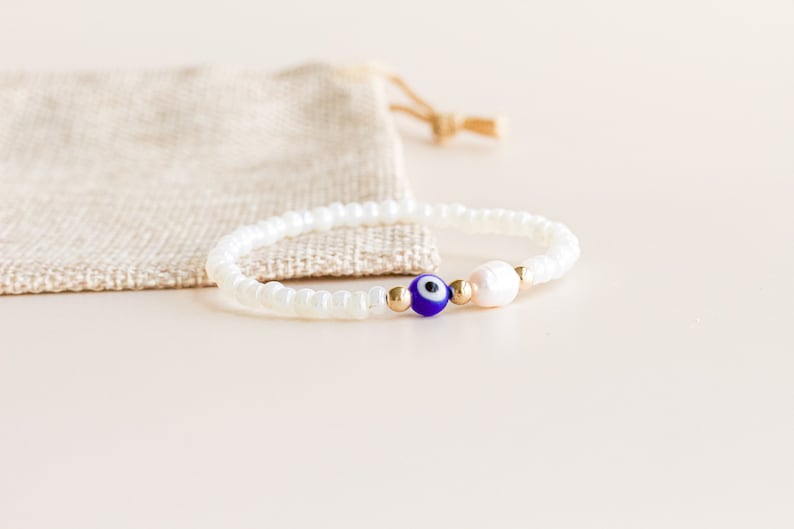 Peut inclure: Un bracelet compos&eacute; de petites perles blanches opaques, d'une perle unique en forme d'&oelig;il bleu et blanc, d'une petite perle d'eau douce blanche et de deux minuscules perles intercalaires m&eacute;talliques dor&eacute;es sur un cordon &eacute;lastique fin.