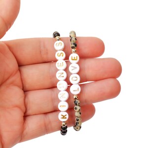 May include: Two bracelets, one with black and white beads and the other with white beads that spell out "LOVE" and "KINDNESS" in gold lettering.