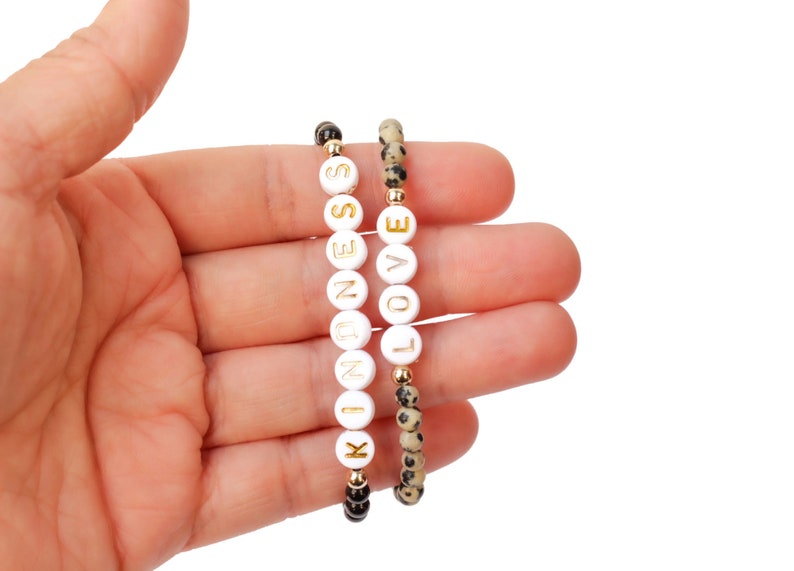 May include: Two bracelets, one with black and white beads and the other with white beads that spell out "LOVE" and "KINDNESS" in gold lettering.