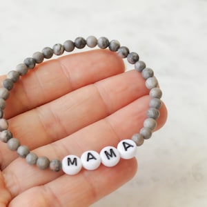 May include: A beaded bracelet featuring small, round, grey and white marbled stone beads. The bracelet includes four white, circular, flat beads with the black printed letters M, A, M, and A, spelling out the word MAMA in the center.