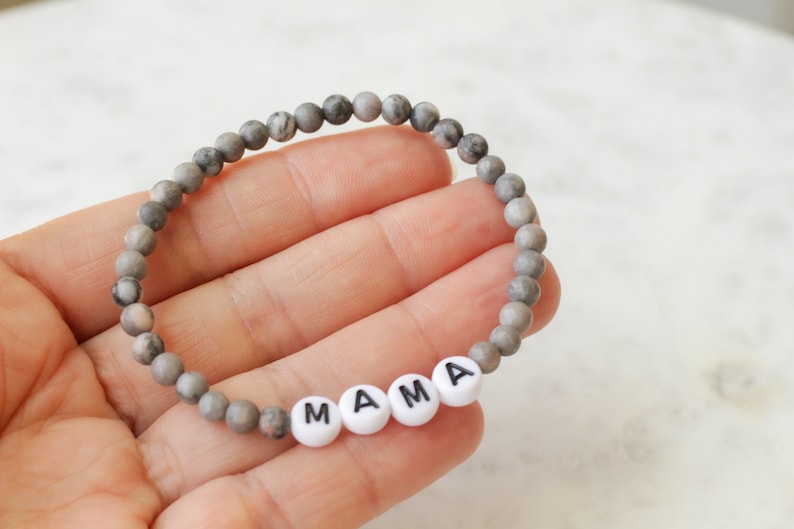 May include: A beaded bracelet featuring small, round, grey and white marbled stone beads. The bracelet includes four white, circular, flat beads with the black printed letters M, A, M, and A, spelling out the word MAMA in the center.