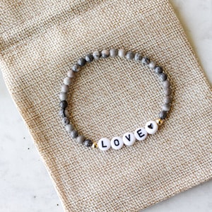 May include: A stretch bracelet featuring small, round, grey and white mottled stone beads. The bracelet includes four white circular beads with black letters spelling out LOVE, followed by a small black heart symbol, accented by gold-tone beads.