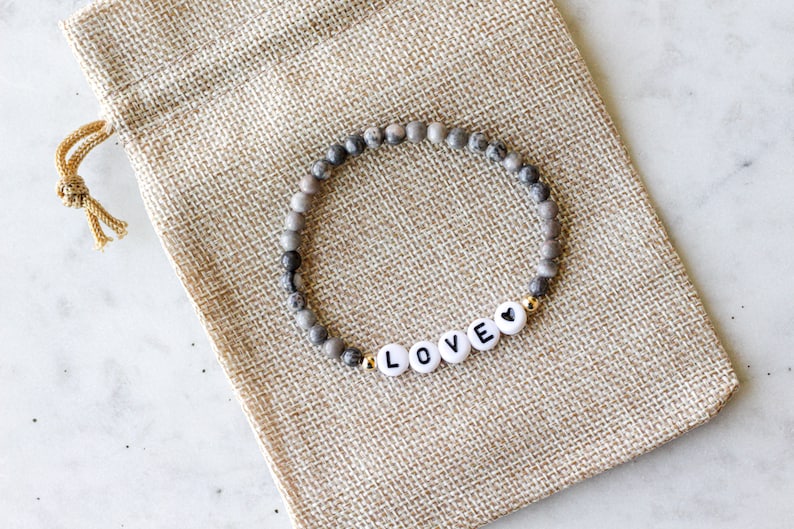May include: A stretch bracelet featuring small, round, grey and white mottled stone beads. The bracelet includes four white circular beads with black letters spelling out LOVE, followed by a small black heart symbol, accented by gold-tone beads.