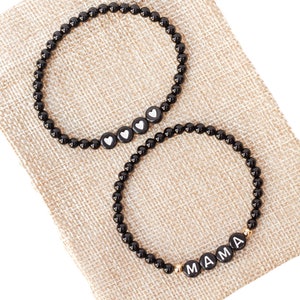 May include: Two black beaded bracelets with gold accents. One bracelet has white hearts and the other says "MAMA".