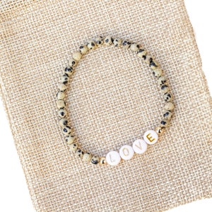 May include: A beige and black speckled stone bracelet with gold accents and white letter beads spelling "LOVE". The bracelet is displayed on a beige burlap pouch.