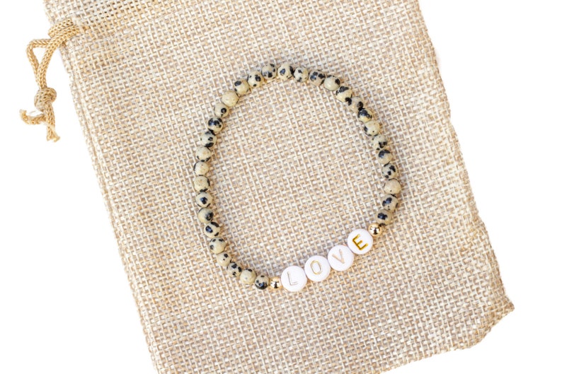 May include: A beige and black speckled stone bracelet with gold accents and white letter beads spelling "LOVE". The bracelet is displayed on a beige burlap pouch.