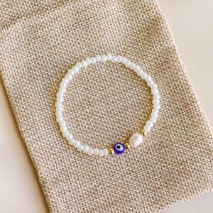 Peut inclure: Un bracelet de perles compos&eacute; de petites perles de verre blanches, d'une perle &oelig;il bleu et blanc, d'une petite perle d'eau douce blanche et de minuscules perles intercalaires m&eacute;talliques dor&eacute;es dispos&eacute;es en cercle sur une pochette en tissu beige.