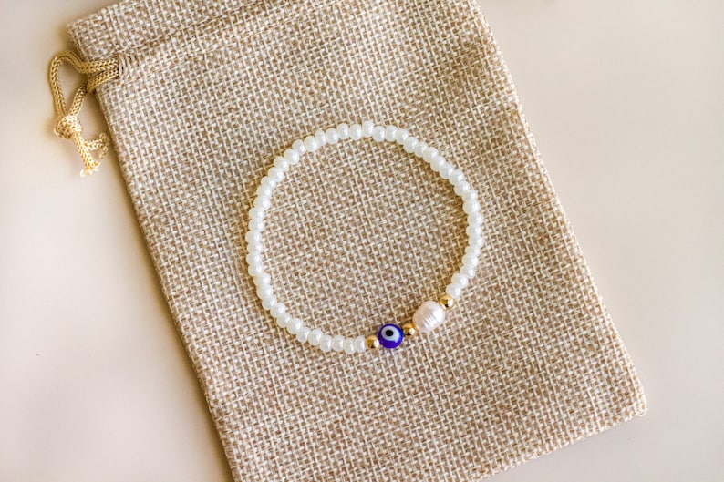 Peut inclure: Un bracelet de perles compos&eacute; de petites perles de verre blanches, d'une perle &oelig;il bleu et blanc, d'une petite perle d'eau douce blanche et de minuscules perles intercalaires m&eacute;talliques dor&eacute;es dispos&eacute;es en cercle sur une pochette en tissu beige.