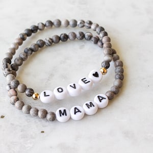 May include: Two stretch bracelets made of grey and white marbled stone beads. One bracelet features white letter beads spelling LOVE with a black heart symbol, and the other features white letter beads spelling MAMA, both accented with small gold beads.