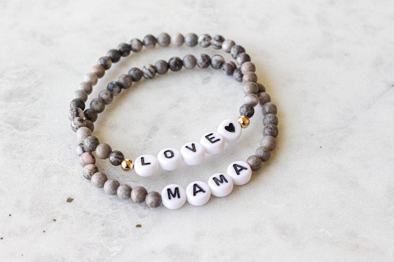 May include: Two stretch bracelets made of grey and white marbled stone beads. One bracelet features white letter beads spelling LOVE with a black heart symbol, and the other features white letter beads spelling MAMA, both accented with small gold beads.