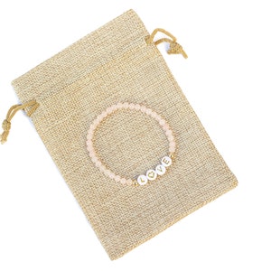May include: A beaded bracelet featuring small, pale pink translucent beads and four white circular letter beads that spell out the word LOVE, with a gold heart symbol replacing the letter O, resting on a tan, textured burlap drawstring pouch.