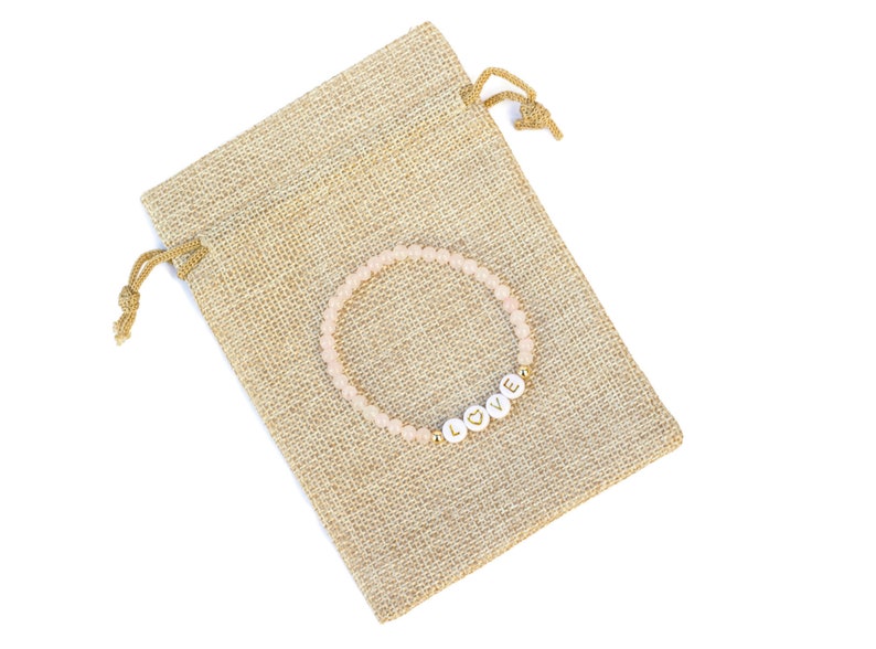 May include: A beaded bracelet featuring small, pale pink translucent beads and four white circular letter beads that spell out the word LOVE, with a gold heart symbol replacing the letter O, resting on a tan, textured burlap drawstring pouch.