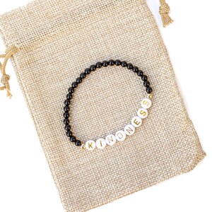May include: A black beaded bracelet with white beads spelling out the word "KINDNESS". The bracelet is on a brown burlap pouch.