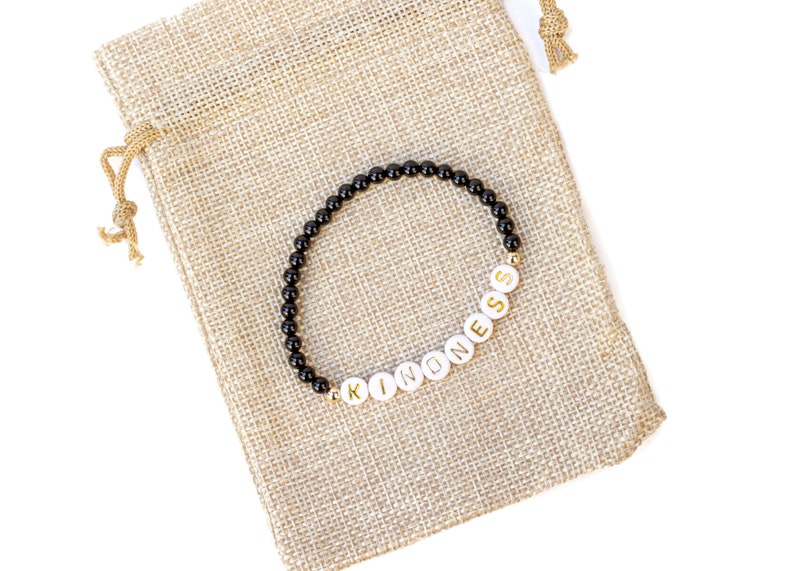 May include: A black beaded bracelet with white beads spelling out the word "KINDNESS". The bracelet is on a brown burlap pouch.