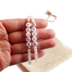 May include: Two beaded bracelets featuring light pink translucent beads and white circular beads with gold lettering. One bracelet spells out MAGGY and the other spells out MAMA with a gold heart symbol. The bracelets are strung on elastic cord.