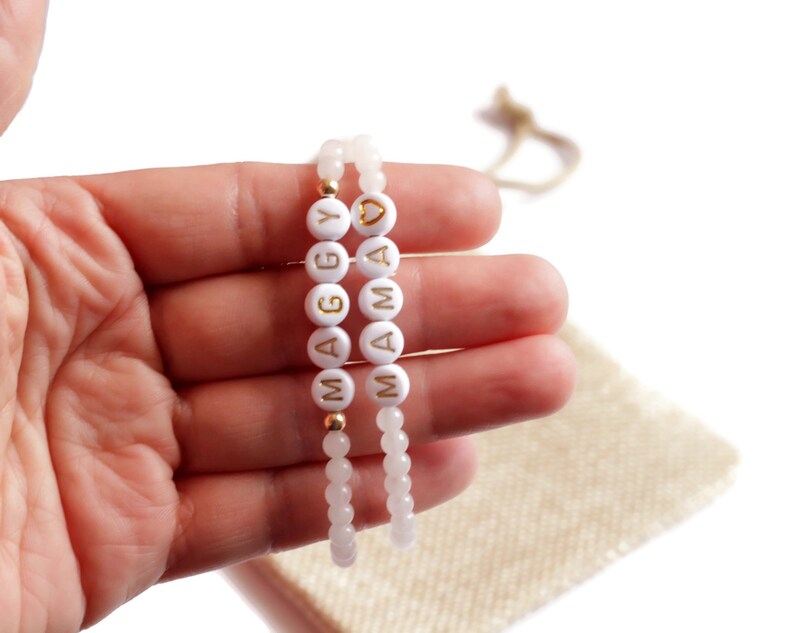 May include: Two beaded bracelets featuring light pink translucent beads and white circular beads with gold lettering. One bracelet spells out MAGGY and the other spells out MAMA with a gold heart symbol. The bracelets are strung on elastic cord.