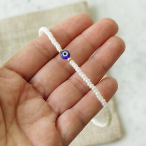 May include: A white beaded bracelet with a blue and white evil eye charm. The bracelet is on a hand.
