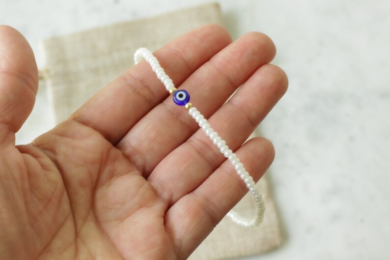 May include: A white beaded bracelet with a blue and white evil eye charm. The bracelet is on a hand.