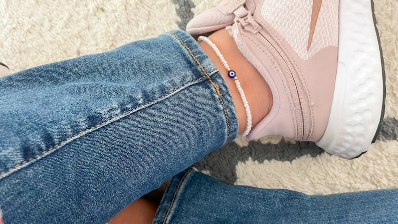 May include: White beaded ankle bracelet with a blue evil eye charm. The bracelet is worn on a person's ankle with blue jeans and pink sneakers.