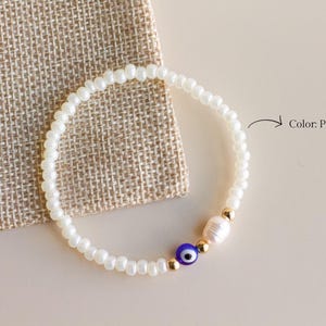 Peut inclure: Un bracelet de perles compos&eacute; de petites perles blanc perle, d'une perle &oelig;il bleu et blanc, d'une perle blanche et de deux perles intercalaires dor&eacute;es. Le texte Color: Pearl White est imprim&eacute; &agrave; c&ocirc;t&eacute; du bracelet avec une fl&egrave;che pointant vers lui.