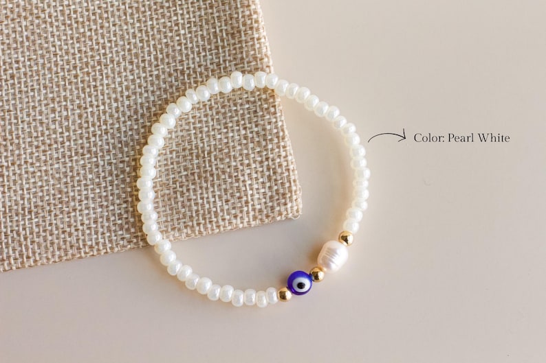 Peut inclure: Un bracelet de perles compos&eacute; de petites perles blanc perle, d'une perle &oelig;il bleu et blanc, d'une perle blanche et de deux perles intercalaires dor&eacute;es. Le texte Color: Pearl White est imprim&eacute; &agrave; c&ocirc;t&eacute; du bracelet avec une fl&egrave;che pointant vers lui.