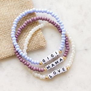 May include: Three beaded bracelets in shades of purple, white, and light blue. The bracelets have white letter beads spelling out "GIGI", "MAMA", and "LOVE". The bracelets are on a marble surface with a burlap sack in the background.