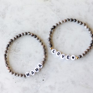 May include: Two beaded bracelets made of small, round, grey and white marbled stone beads. One bracelet features white letter beads spelling MAMA, while the other features white letter beads spelling LOVE followed by a small black heart symbol.